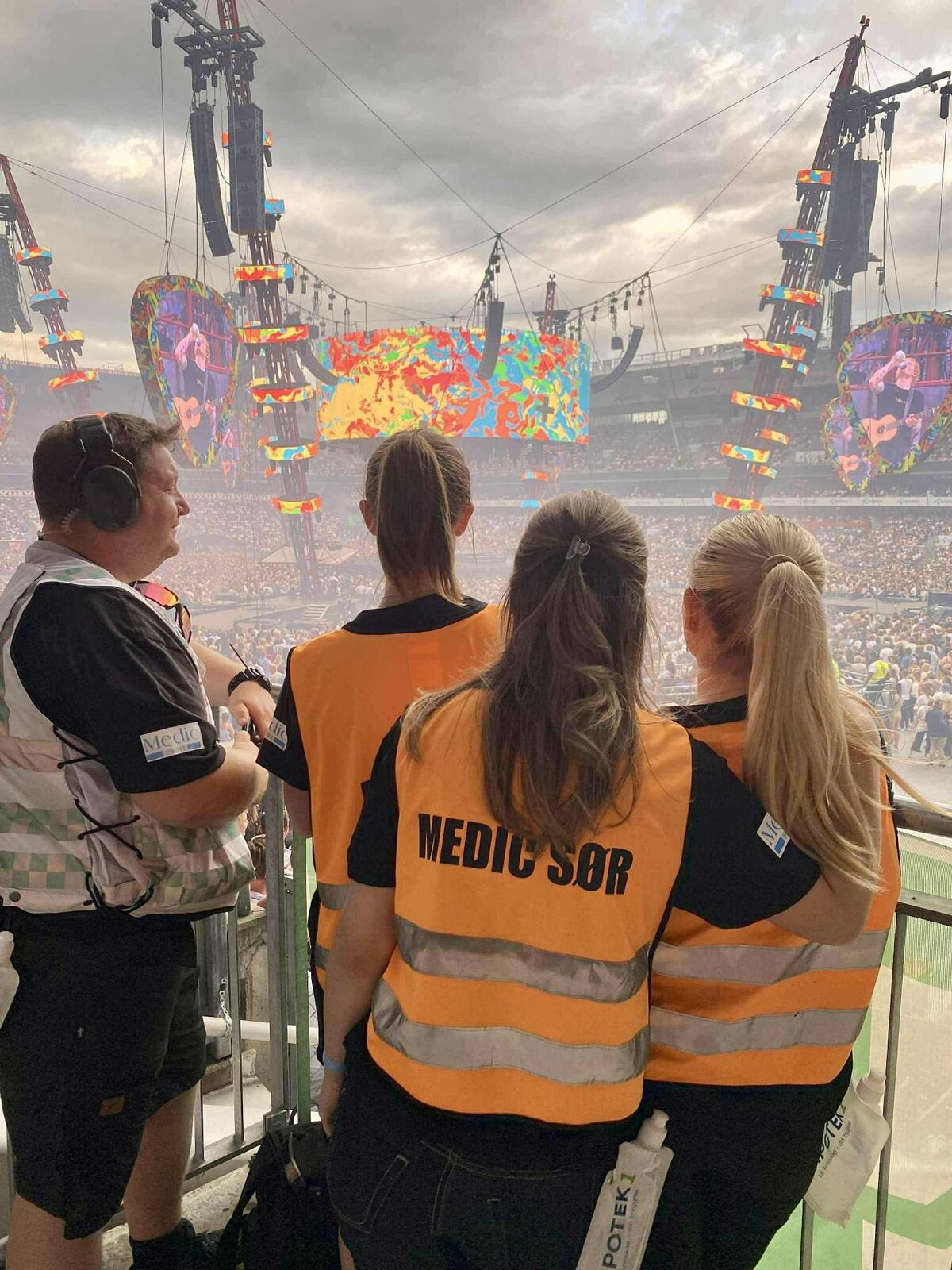 Fire fra Medic Sør i oransje vester står med ryggen til på en tribune og ser utover publikum på en stadionkonsert.